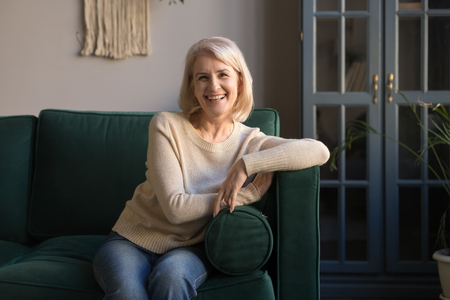 Portrait of smiling grey haired mature woman looking at camera, happy middle aged grandmother sitting on comfortable couch at home, single senior positive satisfied lady posing for photo, old beautyの写真素材