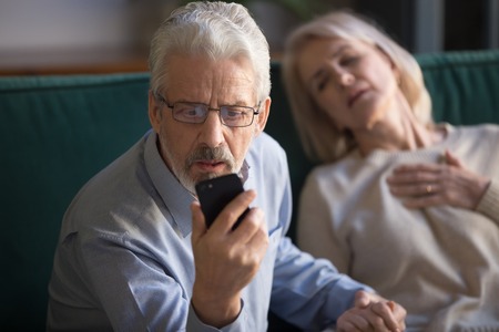 Unhappy mature man, husband calling emergency, holding wife hand, looking on phone screen, woman having heart attack, touching chest, suffering from heartache disease at home, feeling painの写真素材