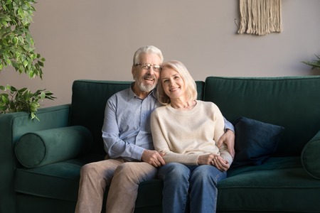 Portrait of hugging aged man and woman sitting on comfortable couch at home, looking at camera, loving grey haired husband embracing smiling beautiful wife, mature family posing for photoの写真素材