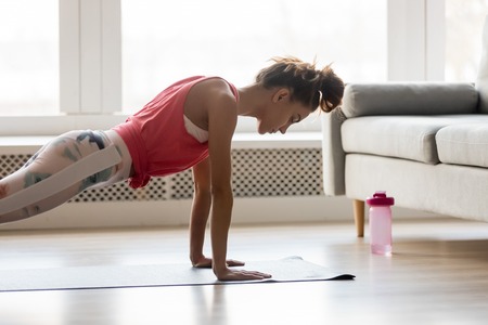 Young attractive sportive woman wearing activewear doing push ups or press ups exercise position on sport mat at home, practicing yoga plank pose, working out indoors, healthy active lifestyle conceptの写真素材