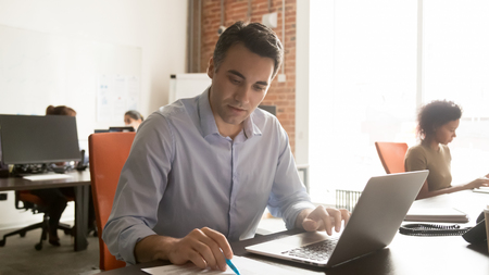 Focused male worker sit at desk in coworking space check data in document working at laptop, concentrated man employee analyzing paperwork busy typing at computer in shared officeの写真素材
