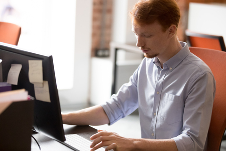 Focused redhead millennial man sit at table work at computer consulting clients online, concentrated red-haired young male employee type email to client customer on pc, busy worker in coworking spaceの写真素材