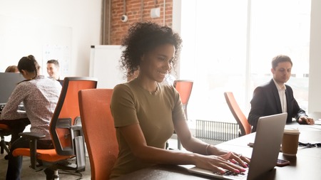 Smiling african American millennial employee sit at desk working on laptop consult client online in shared office, happy positive black woman worker busy typing email to customer in coworking spaceの写真素材