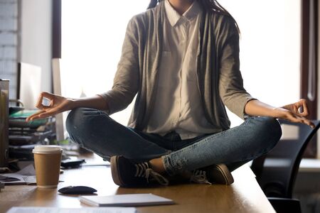 Cropped close up of female sitting at office desk in lotus position do yoga exercise. Woman employee meditating during working day. Stress relief, good healthy habit, self-control and balance conceptの写真素材