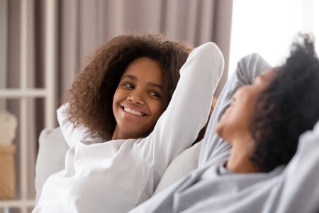Close up smiling African American teen daughter relaxing with mother, leaning back with hands behind head on cozy sofa, happy mum and teenage girl talking, chatting, having fun at home togetherの写真素材