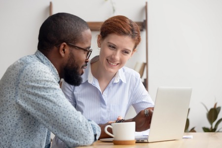 Two diverse smiling businesspeople looking at laptop screen sitting at desk discussing online project, startup, sharing ideas, analyzing results, brainstorming together, thinking of business strategyの写真素材