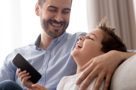 Close up of smiling young father spend time relax with excited preschooler son rest on couch using smartphone, overjoyed dad and boy child laugh play or watch cartoon on cellphone at homeの写真素材