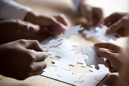 Close up members of multiracial team assembling white puzzle, African American and Caucasian people...