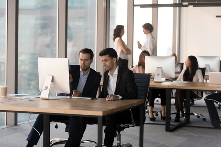 Focused millennial men sit at pc working together discussing project in shared workspace, diverse businesspeople busy using computers, preparing reports in coworking office. Cooperation conceptの写真素材