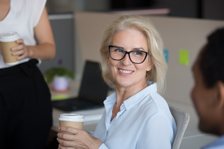 Close up smiling mature businesswoman listening to colleague during coffee break in office, employees having pleasant conversation, discussing funny news, enjoying free time at work togetherの写真素材