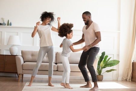 Happy funny active african family with cute little kid daughter dancing at home, carefree cheerful black parents mom dad and small child girl having fun jumping laughing enjoy leisure in the morningの写真素材