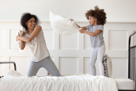 Happy african american family mixed race mom and little cute daughter having fun pillow fight on bed, smiling single mother laugh play funny game with small active child girl in bedroom on leisureの写真素材