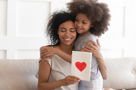 Happy young african american single mom embracing little cute child daughter thanking for gift, smiling black mum hugging small kid holding greeting card with red heart bonding on mothers day conceptの写真素材