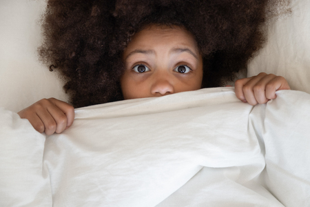Scared cute little african kid girl looking at camera lying in bed cover with blanket, surprised small black child peeking from duvet wake from bad dream sleep feel fear afraid of nightmare, top viewの写真素材