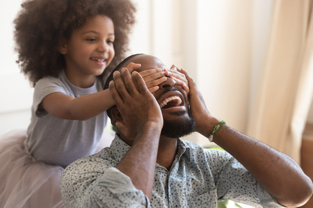Making surprise for daddy concept, cute little african american kid daughter close eyes of excited happy black dad on fathers day, funny small child girl having fun with papa laughing bonding at homeの写真素材