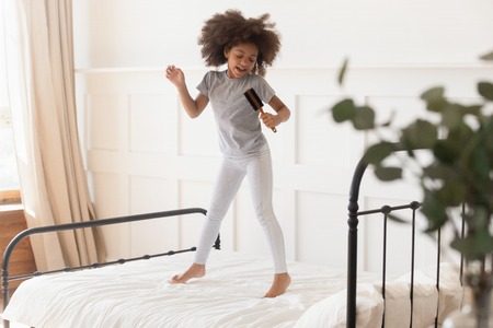 Happy funny cute little african american kid girl jumping on bed singing in microphone hairbrush at home, small mixed race child having fun dancing to music playing in bedroom alone in the morningの写真素材