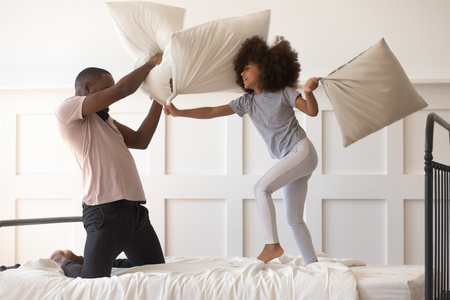 Cute active kid daughter playing pillow fight on bed with african american dad, happy mixed race father and funny little child girl having fun in bedroom black family enjoy leisure morning activityの写真素材