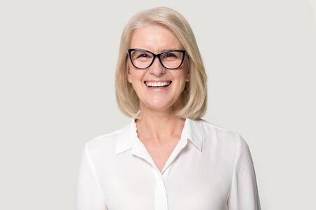 Head shot portrait laughing old businesswoman in glasses white blouse looks at camera feels happy pose isolated on grey studio background, experienced professional business coach teacher concept image