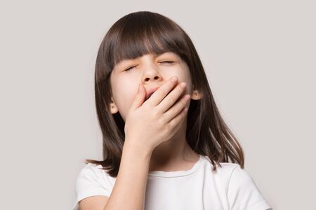 Little brown-haired girl feeling tired or sleepy cover mouth with hand closed eyes yawning isolated on beige studio background, close up face adorable preschool kid suffer from boredom concept imageの写真素材
