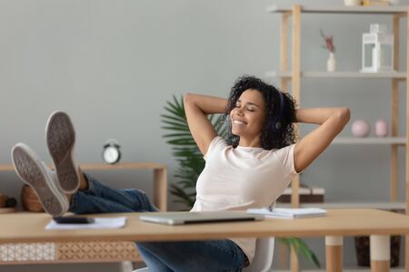 Happy relaxed african woman wear wireless headphones listening to lounge music relaxing after work hands behind head, calm serene black girl student feel stress free relief sit at home office deskの写真素材