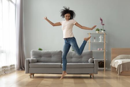 Carefree joyful african american millennial girl jumping dancing alone at home, happy funky active young fit mixed race woman having fun laughing flying in air in studio apartment room interiorの写真素材