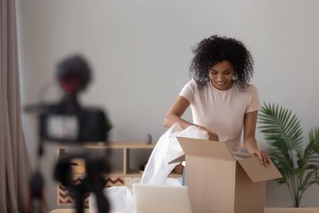 Happy african american young woman vlogger open parcel shooting video blog on camera make review on internet shopping order at home, smiling black girl blogger recording vlog for online channelの写真素材