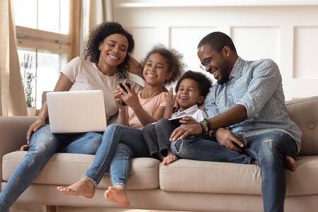 Cheerful african parents and kids laugh use devices together sit on sofa, tech addicted family with children hold laptop phone digital tablet having fun with gadgets at home, technology dependenceの写真素材