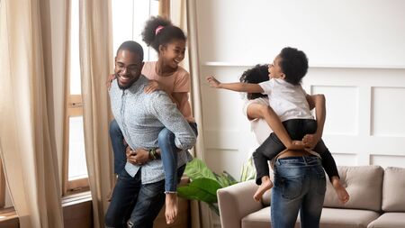 Happy african family mom and dad piggyback little children laugh play in living room, smiling black parents carrying cute kids on back having fun enjoy leisure active game together feel joy at homeの写真素材