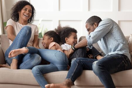 Happy cheerful african american family with cute little kids tickle laugh sit on sofa together,...