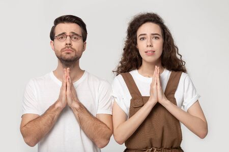 Young couple folding hands together having pleading look praying posing isolated on grey studio...