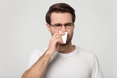 Sick unhealthy young man holding handkerchief paper tissue sneezing wiping blowing runny nose, suffers from unpleasant smell on grey background head shot, concept of seasonal allergy or awful scentの写真素材