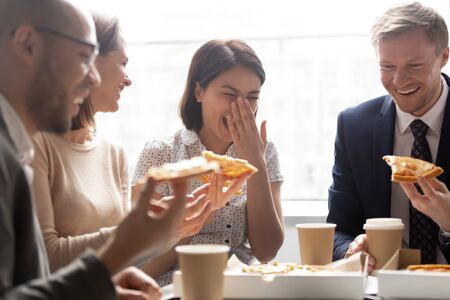 Happy asian worker laugh hold slice eating pizza with cheerful colleagues, smiling multicultural...