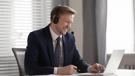 Smiling business man wear suit wireless headset video calling looking at laptop, happy executive communicate in business conference chat on computer consult client online watching webinar in officeの写真素材