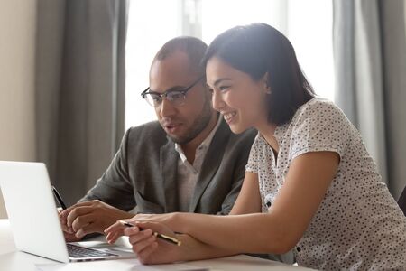 Happy diverse coworkers discuss online work results sit at office desk using laptop, male african...