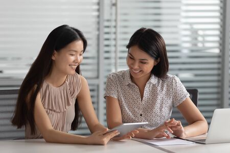 Smiling female korean mentor manager helping asian colleague intern client using digital tablet showing online presentation teaching new worker trainee discuss online project in office, apprenticeの写真素材