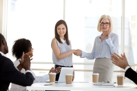 Mature businesswoman shaking hand of female employee at company briefing, middle-aged boss congratulate subordinate with promotion or employment, diverse team colleagues applauding successful internの写真素材