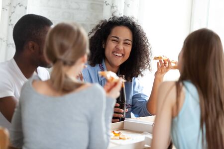 Happy mixed race young woman sitting at table, laughing at friends joke, holding slice of pizza from box, drinking beer, meeting with group of diverse students, enjoying spending free time together.の写真素材