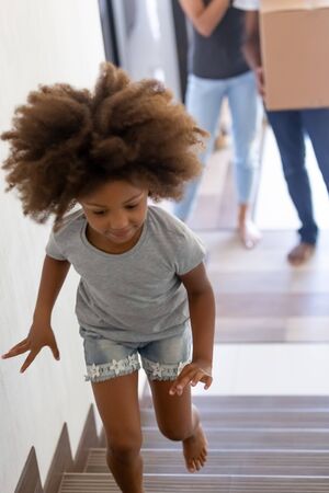 Happy african daughter running to second floor of new two story home parents stand on first below...