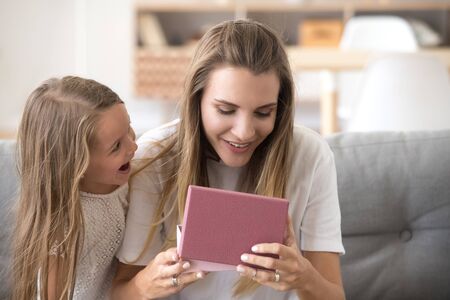 Female and little preschool pretty girl sitting together on couch at home. Surprised happy mother...