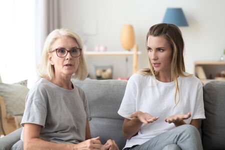 Middle aged woman sitting on couch in living room with young female at home. Serious mother and young daughter family people discussing arguing talking together indoors having unpleasant conversationの写真素材