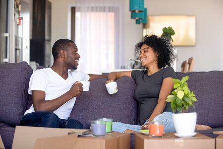 Happy afro couple rest on sofa in living room talking planning future life holds cups drinking tea, heap carton boxes with stuff. Moving day new home, preparations for relocation or renovation conceptの写真素材