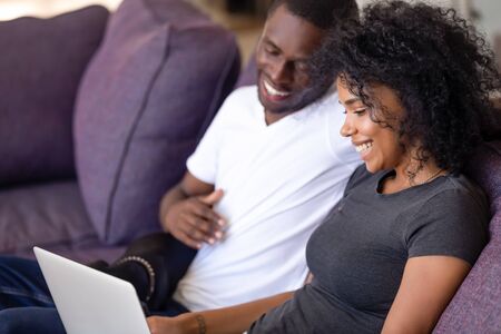 Cheerful african couple sitting on couch resting spending free time watching comedy movie funny videos online at home booking hotel planning vacation modern technology users leisure activities concept.の写真素材