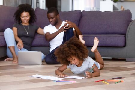 Black family spend free time together in living room at new modern cozy home, couple sitting using laptop watching movie little kid having fun lying on warm wooden floor drawing with colourful pencilsの写真素材