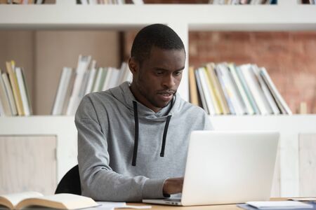 Serious african American male student sit at office desk working typing on laptop studying at home, concentrated black guy busy preparing for exam, doing school assignment or task readyの写真素材