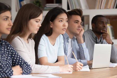Attentive multicultural diverse young people sit at desk listen to teacher talk giving lecture or explaining material, concentrated multiethnic students make noted write down during lesson at collegeの写真素材