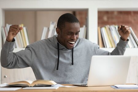 Overjoyed black male student sit at desk look at laptop screen scream passing exam getting high...