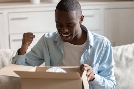 African millennial guy sitting on couch at home looking inside of carton box pleased with received parcel, reliable trusted transportation company, fast easy and quick service of ordered good conceptの写真素材