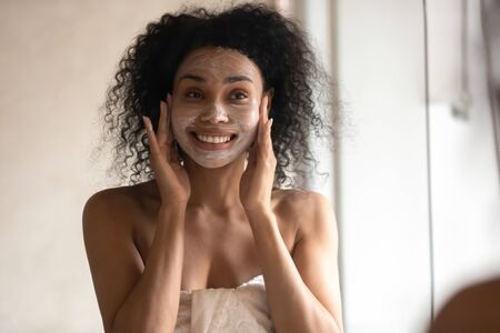 African 30s young woman after shower is wrapped in towel looking in mirror pampering herself applying face mask use anti-aging beauty product feeling happy, skin care, everyday morning routine conceptの写真素材