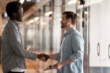 Two diverse male colleagues greeting handshaking talking standing in office hall, smiling african...