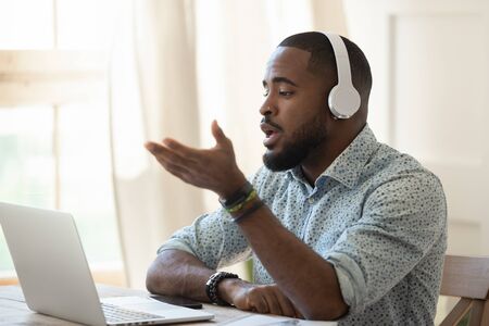 Serious african male interpreter video call teacher wearing headphones looking at laptop talking by...